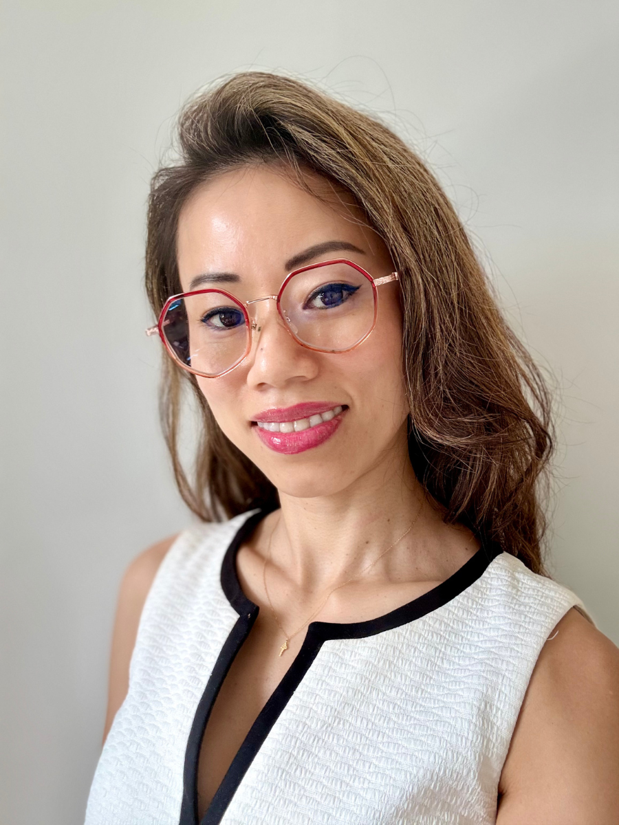 Ms. Shan Shan Jing, a smiling woman wearing glasses is standing in front of a white background, wearing a white sleeveless top